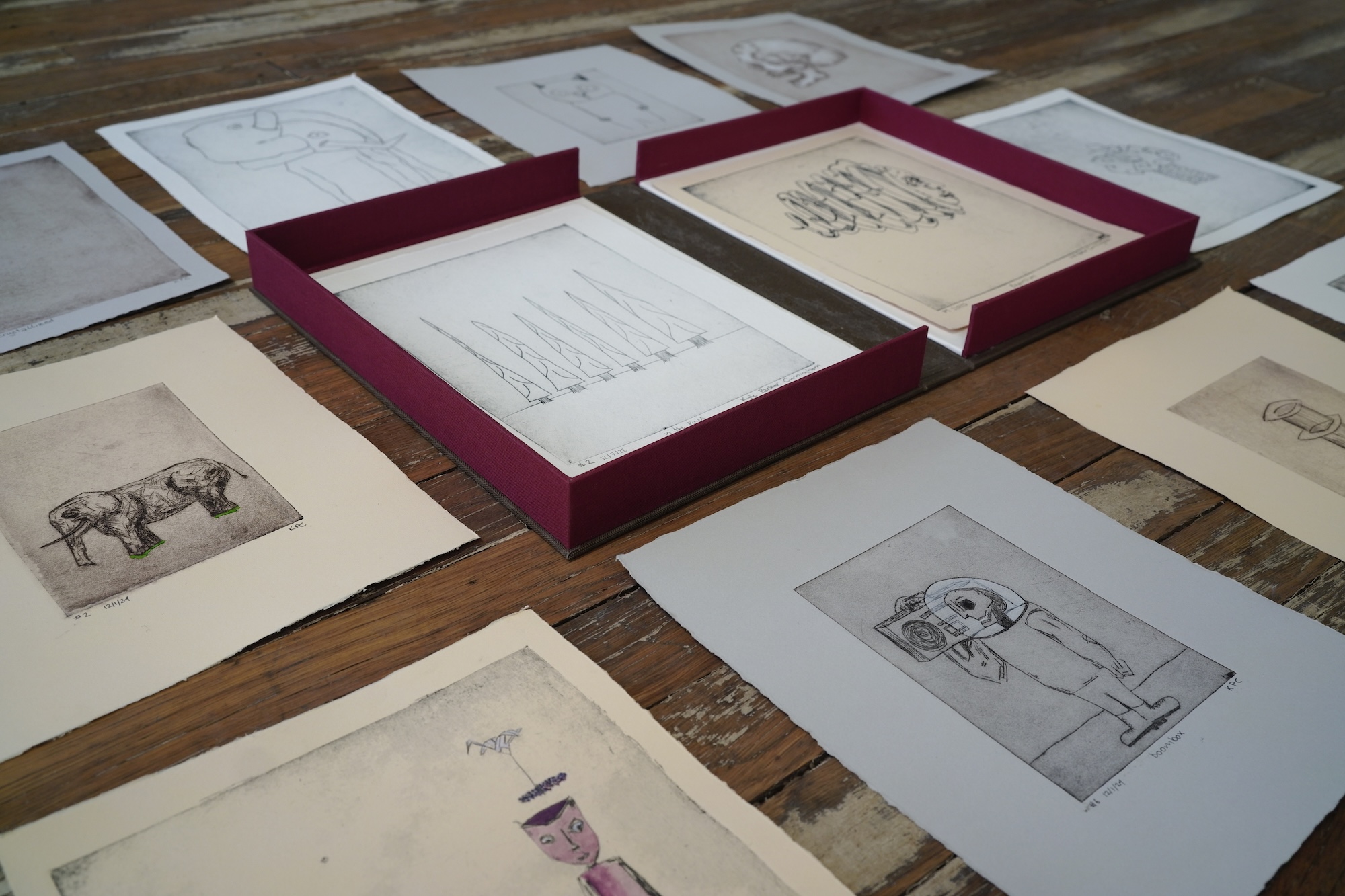 Intaglio prints laid out on a wooden table around a deep red presentation box